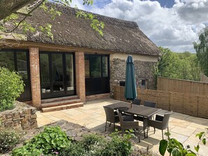 Une maison avec un toit de chaume, des murs en briques et de grandes fenêtres. On y trouve une terrasse avec une table et des chaises, et une clôture en bois en arrière-plan.