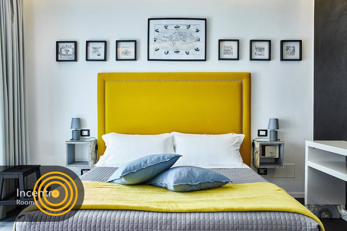 Una habitación de hotel moderna con un cabecero amarillo, una cama con almohadas blancas y azules, y un plaid amarillo. Hay obras de arte enmarcadas en la pared y una mesita de noche con una lámpara a cada lado de la cama.