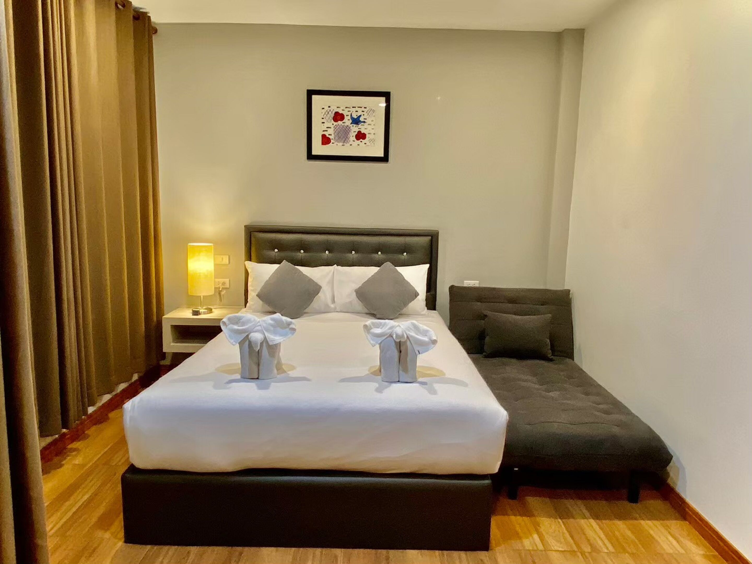 A hotel room with a large bed, a bedside table with a lamp, a grey sofa, and a framed picture on the wall.