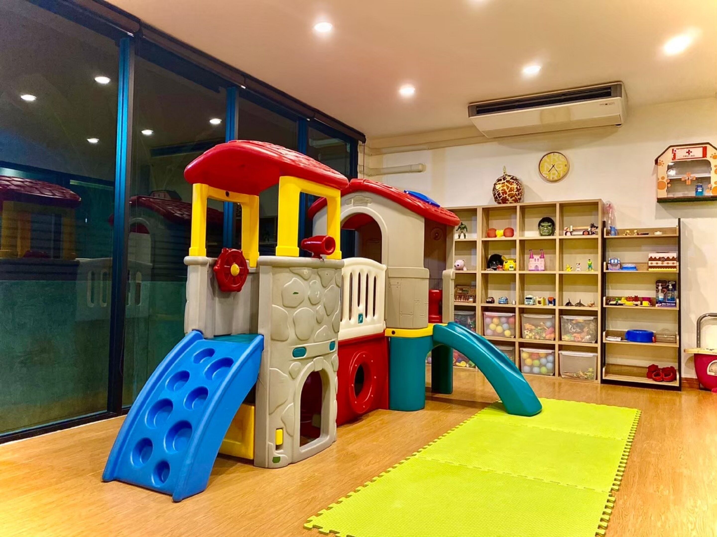 Un espace de jeux pour enfants avec un toboggan et une structure d’escalade, entouré d’étagères remplies de jouets et d’une horloge au mur.