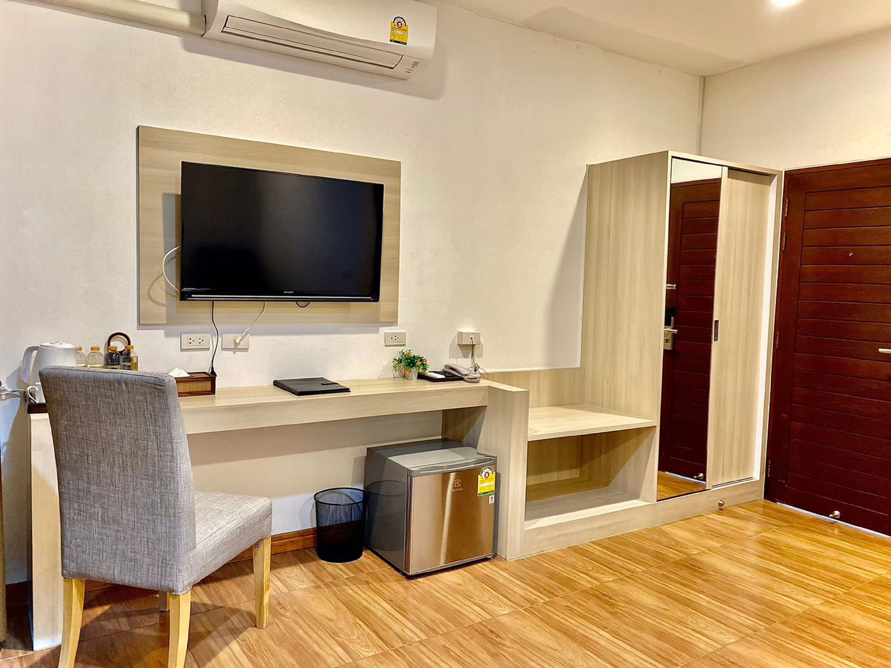 Una habitación moderna con un escritorio de madera, una silla gris, un televisor de pantalla plana en la pared, un cubo de basura pequeño y un armario de madera con espejo.