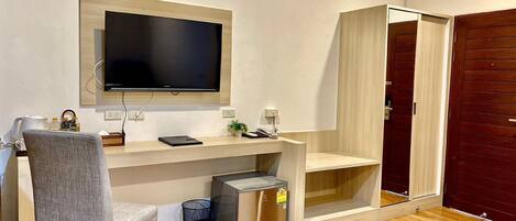 Una habitación moderna con un escritorio de madera, una silla gris, un televisor de pantalla plana en la pared, un cubo de basura pequeño y un armario de madera con espejo.