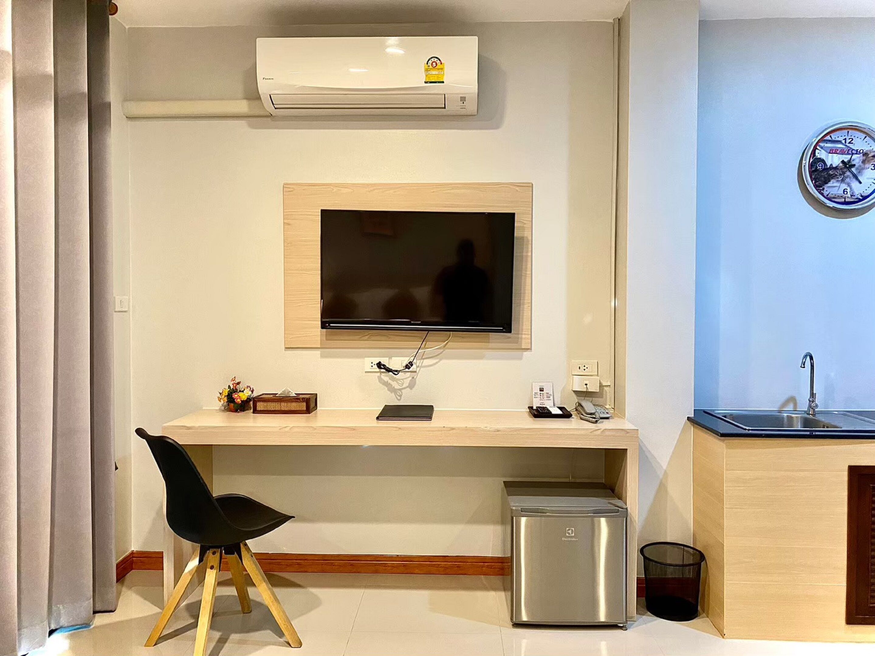 Habitación de hotel compacta con televisor de pantalla plana en la pared, un refrigerador pequeño, escritorio con silla, lavamanos y un reloj en la pared.