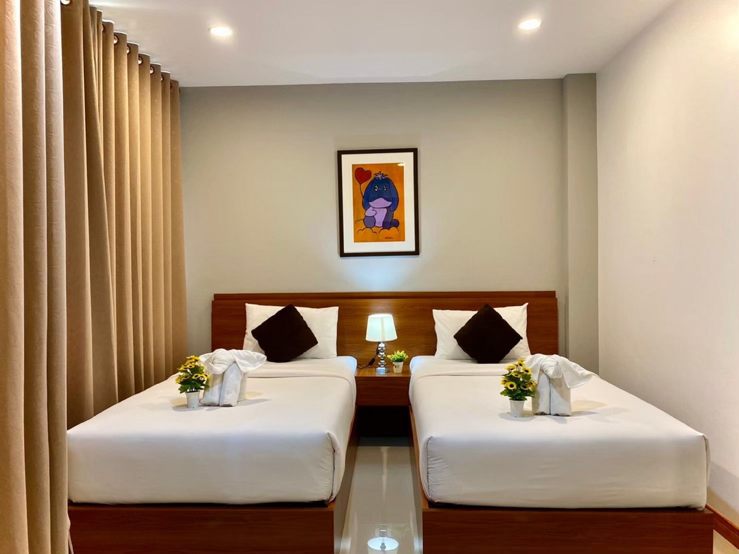 A hotel room with two beds, a wooden headboard, a framed picture on the wall, and a bedside table with a lamp and flowers.