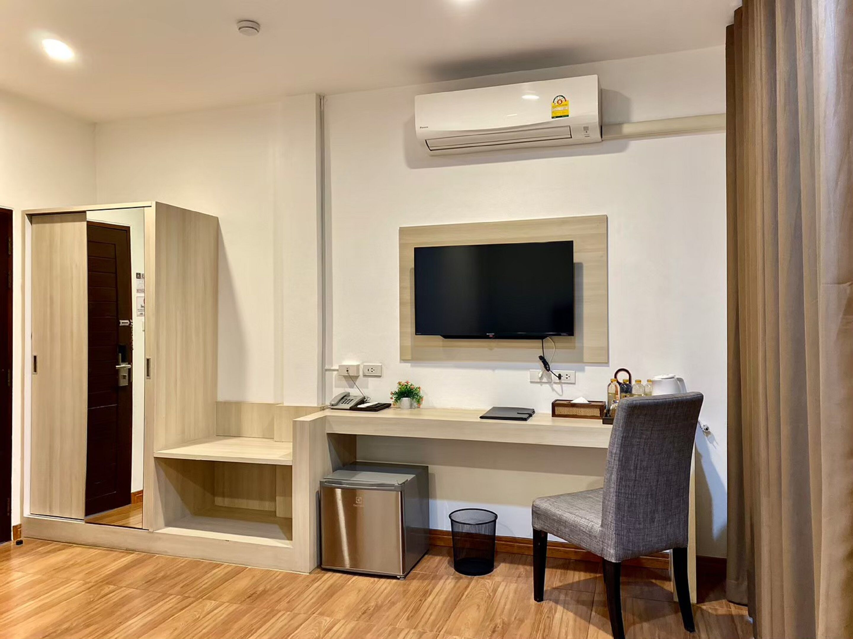 Una habitación de hotel moderna con piso de madera, un escritorio con televisión de pantalla plana, un refrigerador pequeño, un cesto de basura y una silla.