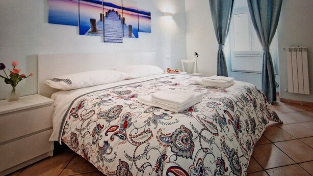 A neatly made bed with a patterned bedspread, a bedside table with a vase of flowers, and a wall-mounted artwork depicting a sunset over a pier. - CASA MINERVA: The Best apartment near Coloseum 5☆ (Roma)