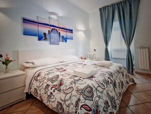 Una cama bien hecha con una colcha estampada, una mesita de noche con un florero y una obra de arte en la pared que muestra un atardecer sobre un muelle.