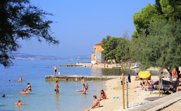 A beach with people swimming and sunbathing, a small building, and trees.