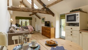 A cozy living room with a sofa, a dining table with a tray of croissants, and a television on a wooden cabinet. - Secret Garden studio at Woodcock Farm (Bristol)