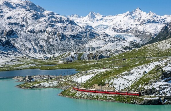 Un train rouge circule sur des voies ferrées qui serpentent autour d’un lac, avec des montagnes enneigées en arrière-plan.