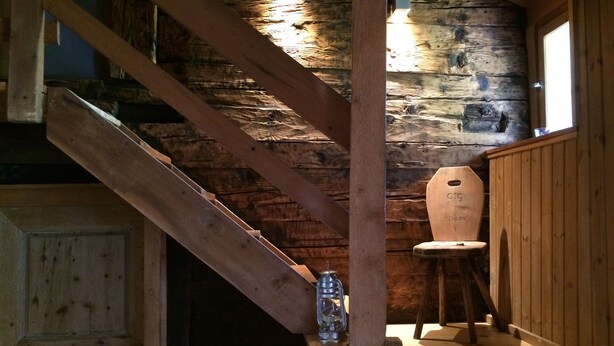 A wooden staircase with a lantern on the landing, a rustic wooden wall, and a wooden chair.