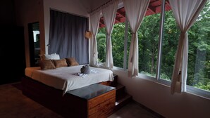 A bedroom with a large bed, a wooden headboard, a window with curtains, and a view of greenery outside.