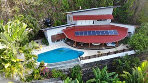 A modern two-story house with a swimming pool, surrounded by lush greenery and a red-roofed deck.