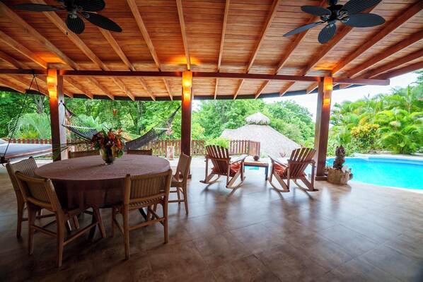 A covered outdoor dining area with a round table, wicker chairs, and a thatched-roof structure in the background. - Private Jungle villa Casa Drop In with private saltwater pool, walk to the beach (CobanSanta Teresa de Cobanoo, Puntarenas)