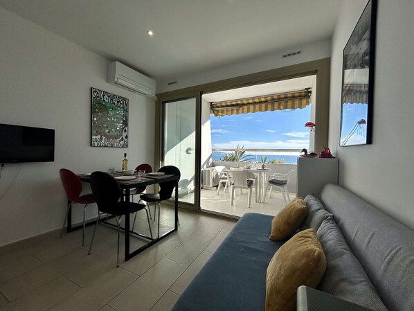 Sala de estar com sofá, mesa de jantar, cadeiras e TV. Possui uma varanda com vista para o mar.