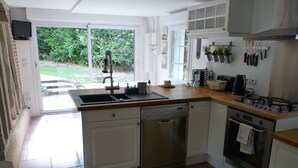 A modern kitchen with a dishwasher, stove, and sink. There is a window with a view of greenery outside. - ---- The cottage --- (ST LEGER DE ROTES)
