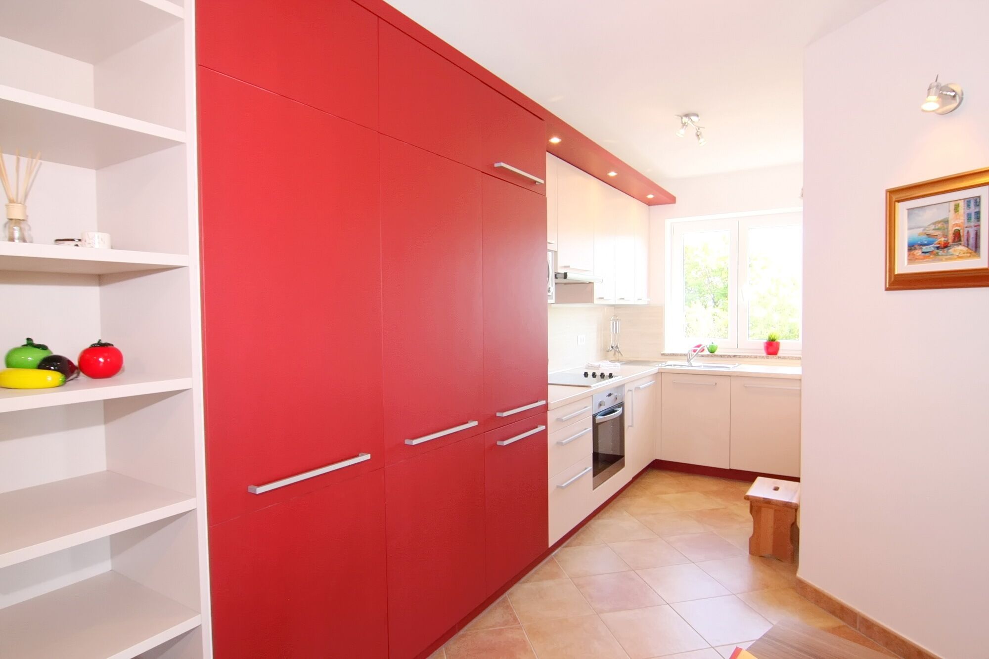 A kitchen with white cabinets, a red cabinet, and a window with a view of greenery.