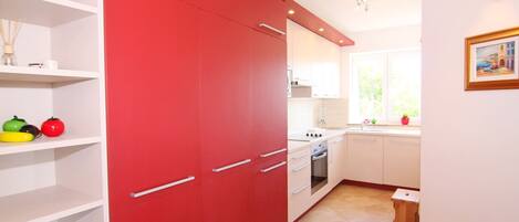 A kitchen with white cabinets, a red cabinet, and a window with a view of greenery.