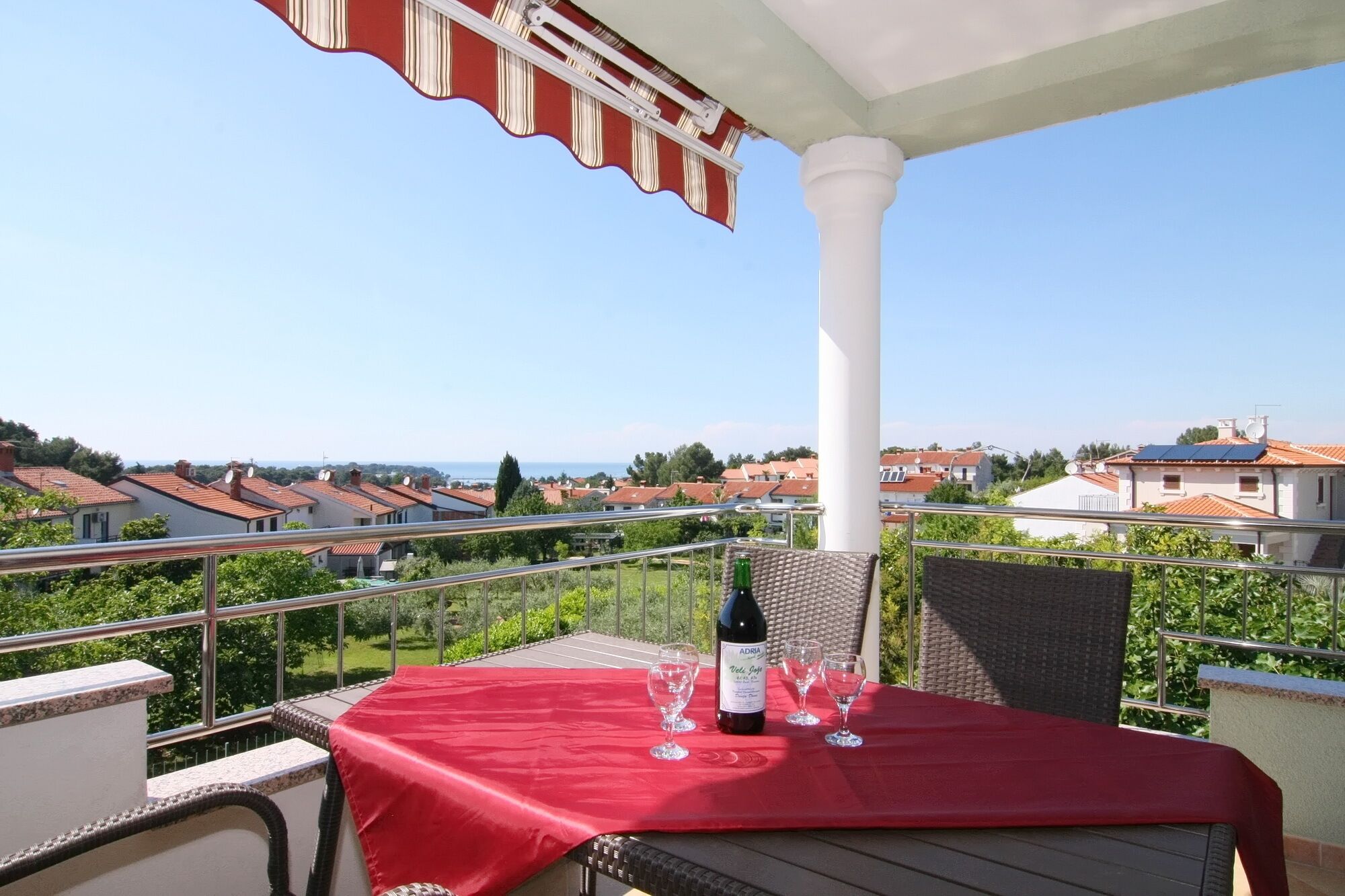 A balcony with a red tablecloth, a bottle of wine, and glasses, overlooking a residential area with red-roofed houses.