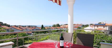 A balcony with a red tablecloth, a bottle of wine, and glasses, overlooking a residential area with red-roofed houses.