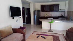 A living room with a sofa, a coffee table, and a TV. There is a kitchen area with cabinets and a refrigerator.