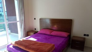 A bedroom with a large bed, purple bedding, a wooden headboard, a nightstand, and a window with curtains.