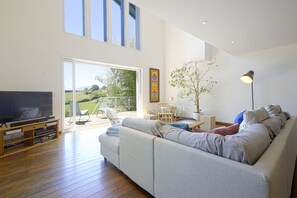 A modern living room with a large sofa, a TV, and a wooden entertainment unit. There is a sliding glass door leading to an outdoor area with a patio chair and a table. - House w/ pool - 12 pers., quiet garden w/ stunning view on the BASQUE COUNTRY (ARCANGUES)