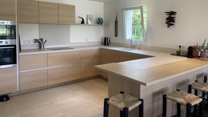 A modern kitchen with light wood cabinets, a large island with bar stools, a built-in oven, and a window providing natural light. - House w/ pool - 12 pers., quiet garden w/ stunning view on the BASQUE COUNTRY (ARCANGUES)