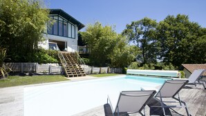 A swimming pool with a wooden deck and lounge chairs, surrounded by trees and a house with a blue exterior. - House w/ pool - 12 pers., quiet garden w/ stunning view on the BASQUE COUNTRY (ARCANGUES)