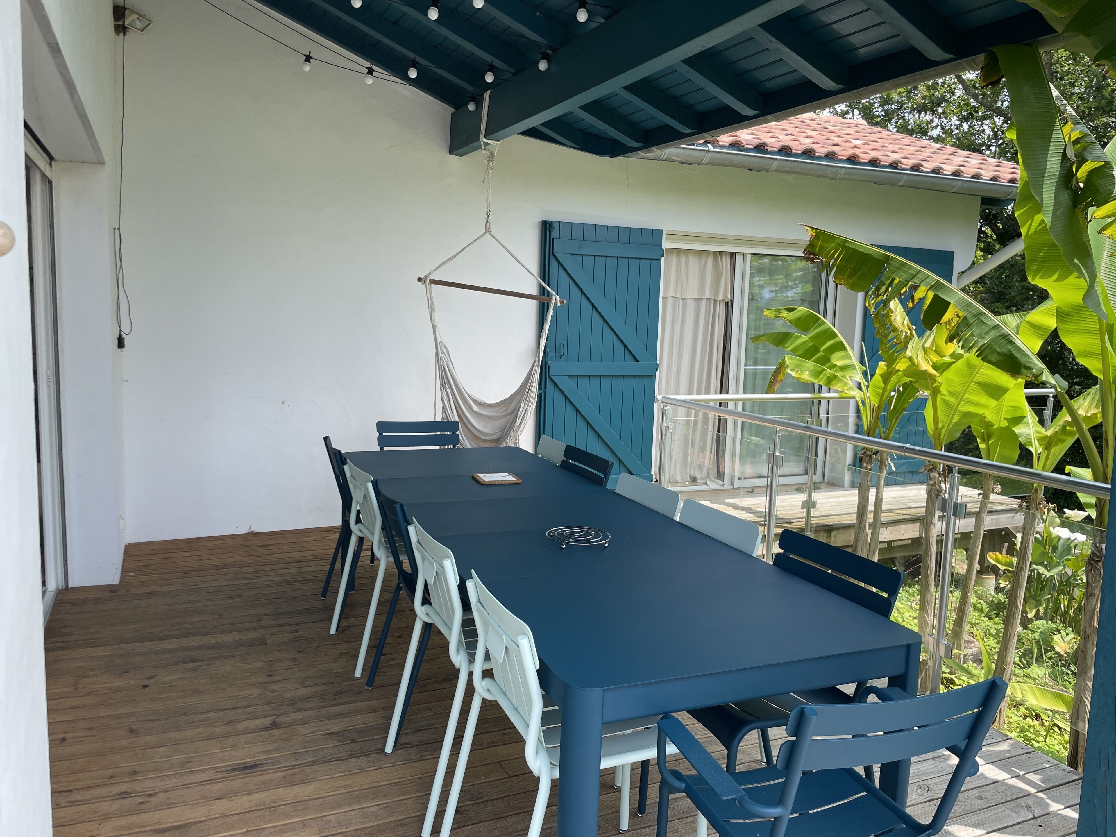 Un espace extérieur abrité, aménagé pour prendre les repas, avec une table et des chaises, un fauteuil suspendu et une vue sur un bâtiment aux volets bleus.