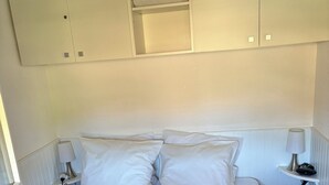 A bed with white bedding, a teal throw, and a white headboard. There is a nightstand with a lamp and a power outlet. Above the bed, there is a white cabinet with a plastic bag inside. - T2 Mezzanine in Ste Maxime Gulf of St Tropez (Sainte-Maxime)