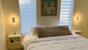 A neatly made bed with a quilted white duvet, two bedside tables with lamps, a small shelf with a plant, and a wall-mounted artwork above the bed. - Romantic, Beautiful Sunsets w/Private Pool (West Punt)