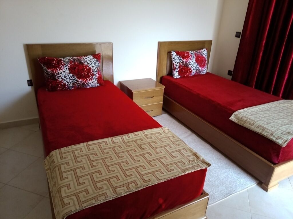 A hotel room with two single beds, each with a red bedspread and matching pillows. There is a wooden nightstand between the beds, and a red curtain on the right side.