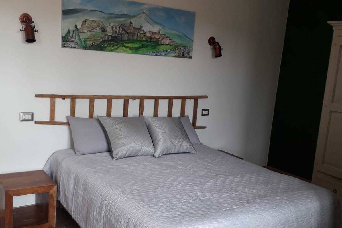 A bedroom with a wooden bed frame, a gray quilted bedspread, two pillows, a nightstand, and a painting of a castle on the wall.