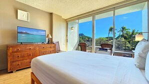 A hotel room with a large bed, a wooden dresser, a flat-screen TV displaying a beach scene, and a balcony with a view of palm trees and mountains.