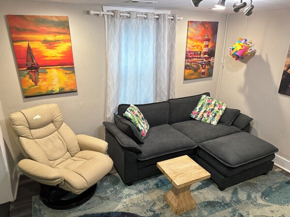 A living room with a beige chair, a dark grey sofa, a small wooden table, and two paintings on the walls.