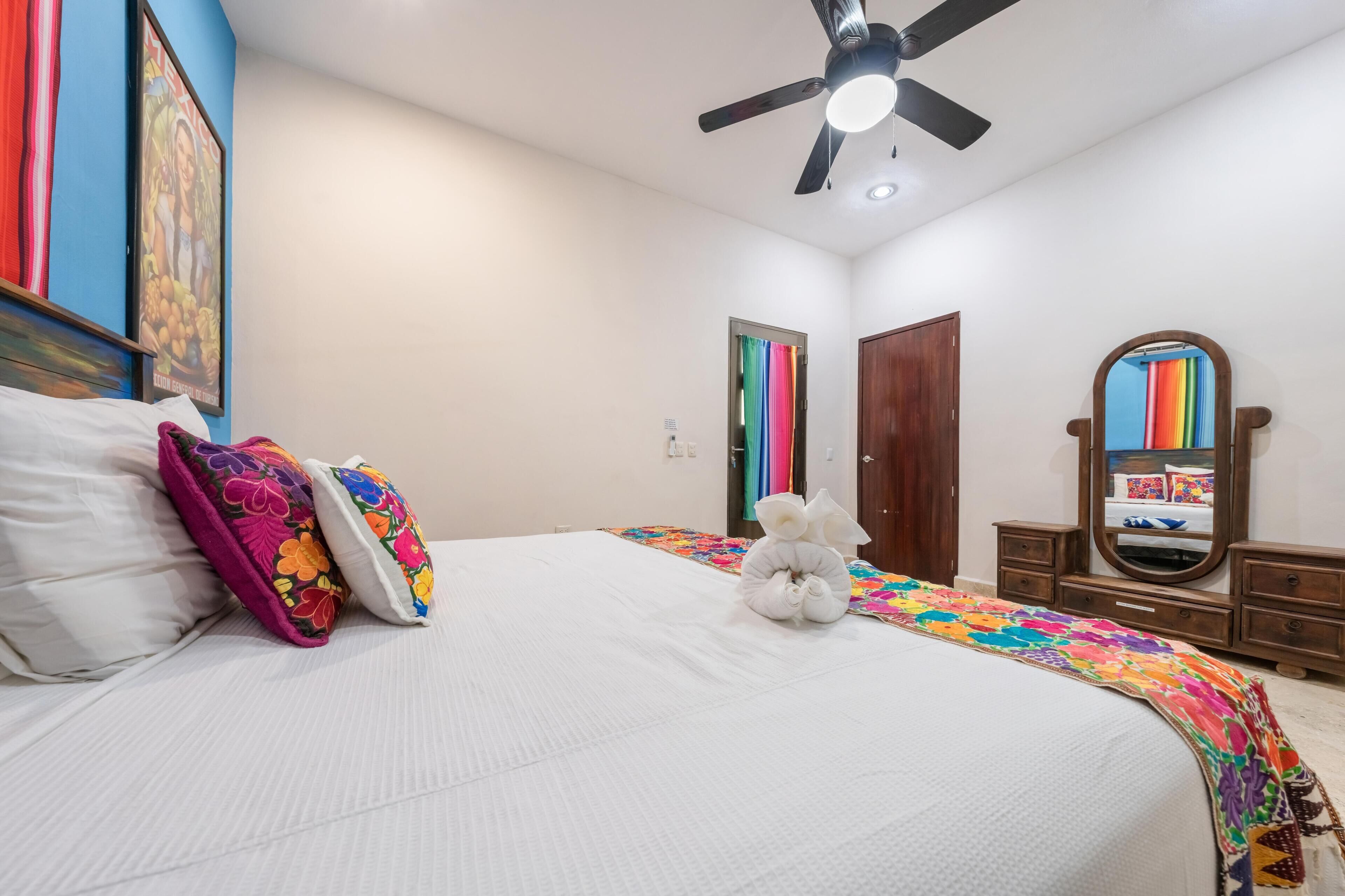 A bedroom with a bed, pillows, a ceiling fan, a dresser, a mirror, and a door.