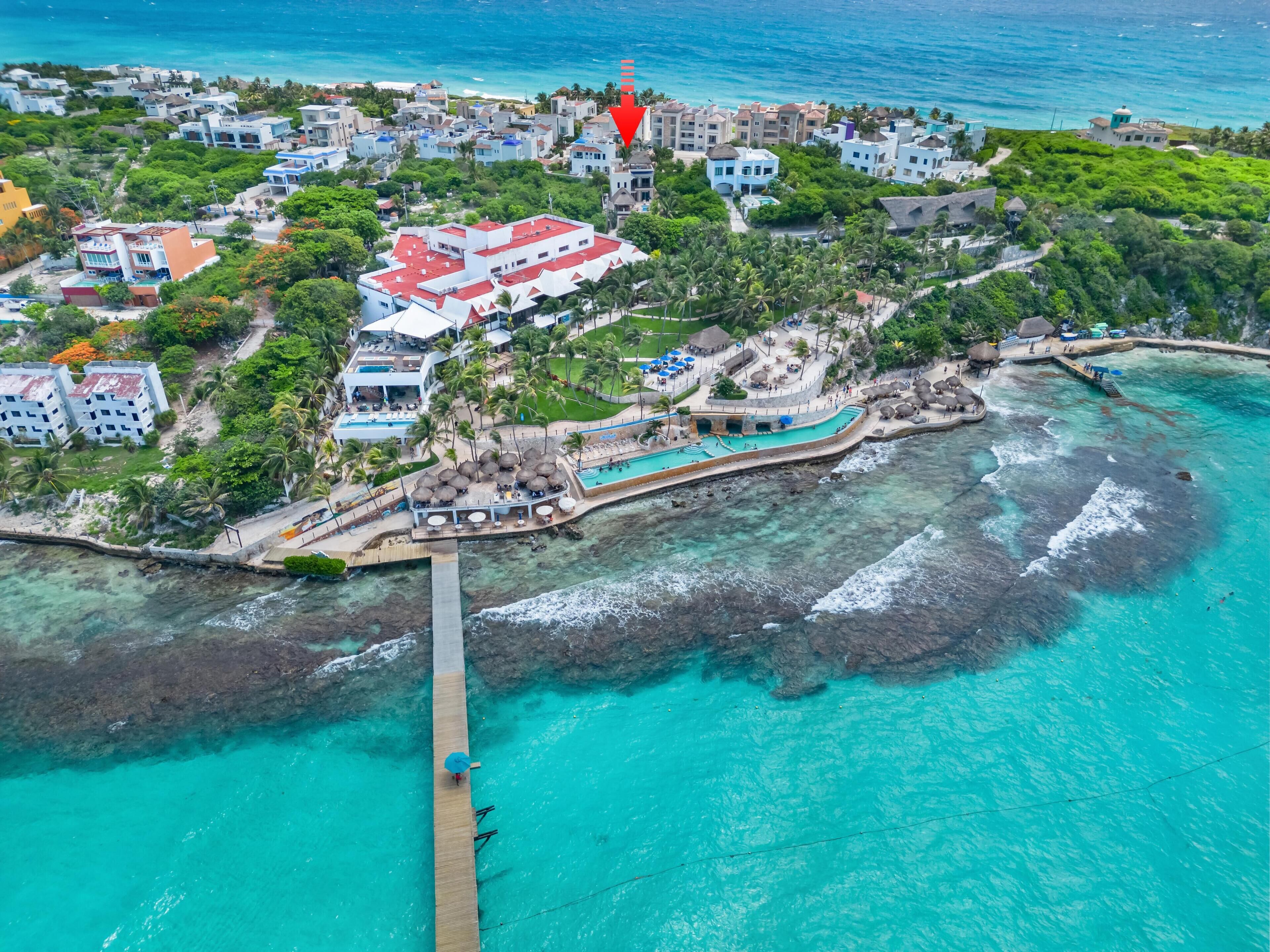 An aerial view of a coastal resort with a pier, clear turquoise waters, and a variety of buildings including a large resort complex with multiple pools and a beach area.
