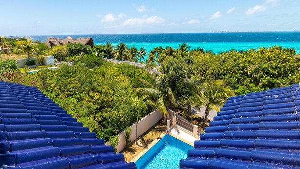 A resort with a swimming pool surrounded by lush greenery and palm trees, overlooking a turquoise ocean.