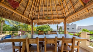An outdoor dining area with a thatched roof, wooden tables and chairs, and a view of greenery and buildings in the background.
