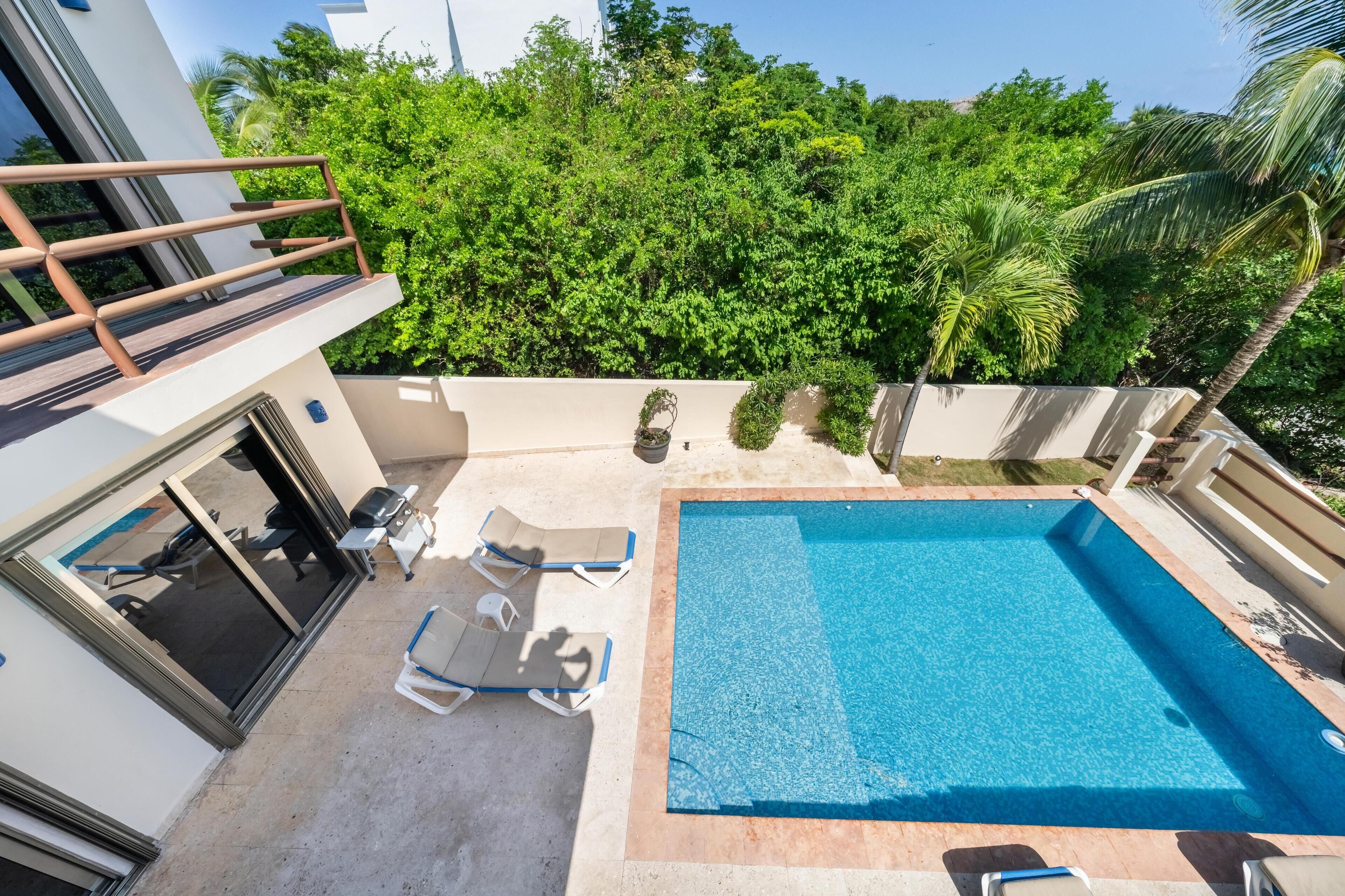 A swimming pool with lounge chairs, a covered seating area, and a view of lush greenery.