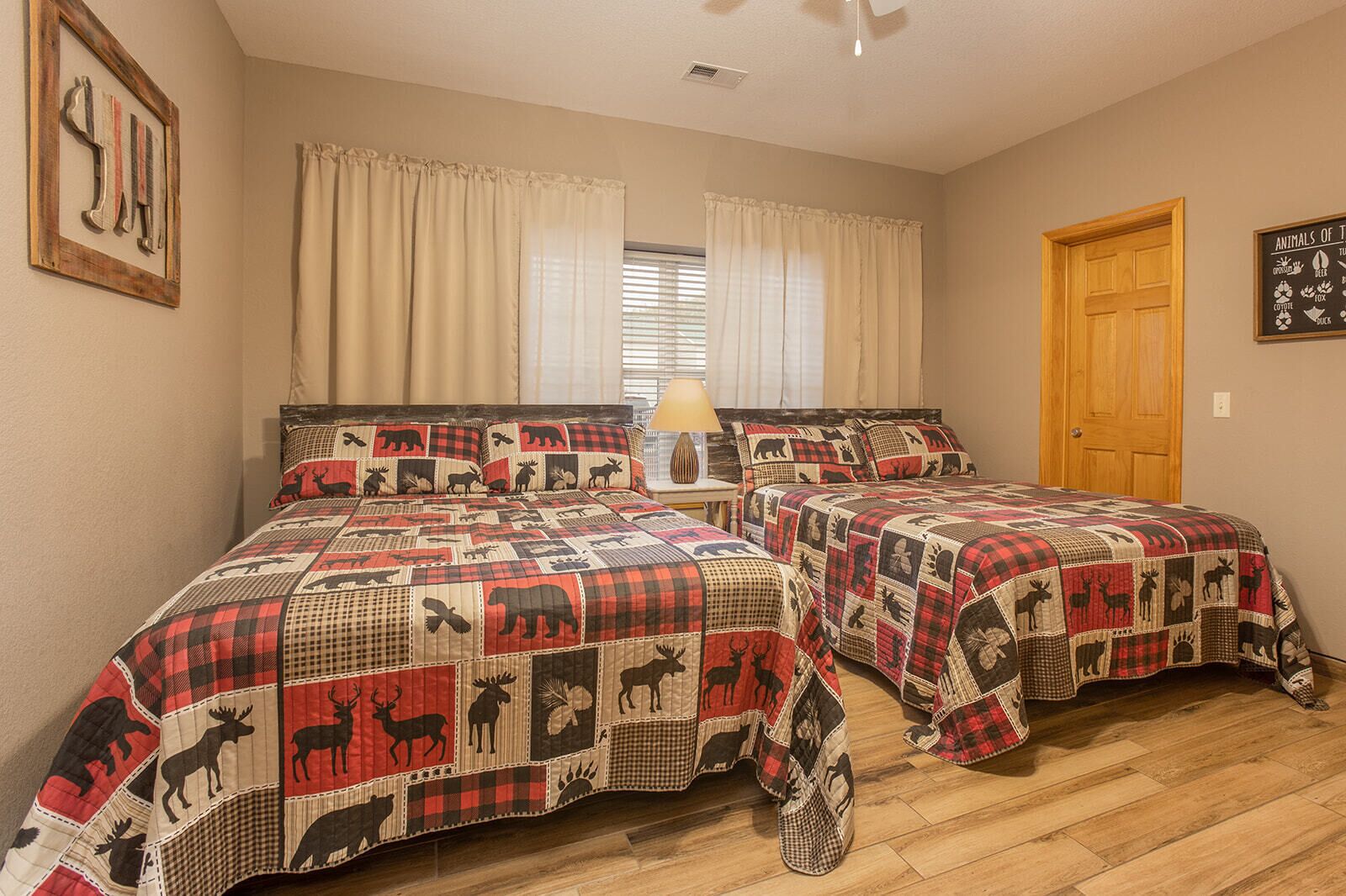 Une chambre avec deux lits, chacun recouvert d’une couverture à carreaux rouges et noirs ornée de motifs d’animaux. On y trouve une porte en bois, une fenêtre avec des rideaux et un cadre photo fixé au mur.