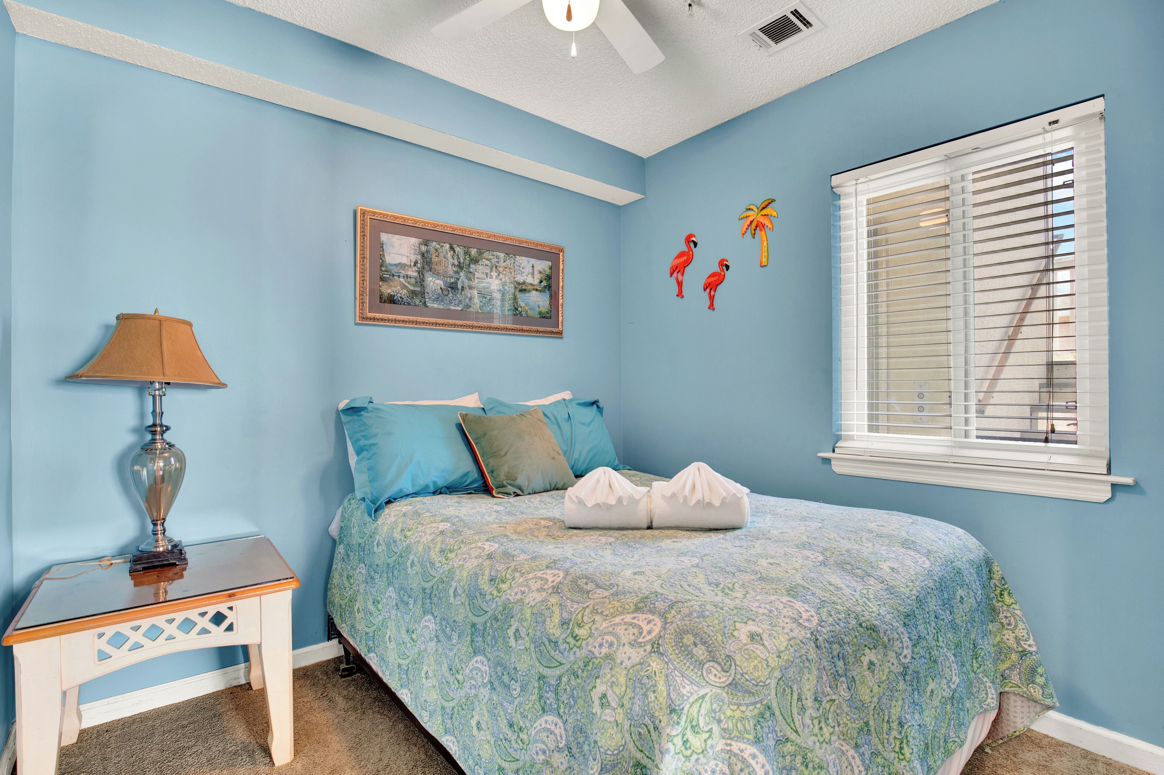 A bedroom with a bed, a nightstand, a lamp, a window with blinds, and wall decorations including a framed picture and wall art.