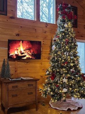 Une pièce chaleureuse avec un décor intérieur en bois, une cheminée, un grand sapin de Noël et un téléviseur.