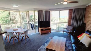 Un amplio salón con una televisión grande sobre un soporte de madera, una mesa de comedor con sillas, un sofá y un balcón con cortinas.