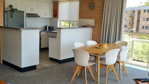 Una cocina compacta con armarios blancos, una encimera de madera y un comedor con mesa y sillas blancas. Se ve un reloj en la pared, y hay una ventana con vistas a edificios.