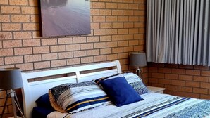 Un dormitorio con pared de ladrillo, cabecera blanca, cama con ropa de cama a rayas, dos lámparas de noche y una ventana con cortinas.