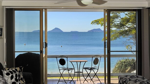 A room with a sliding glass door leading to a balcony with a view of the sea and distant mountains. There is a small table and two chairs on the balcony.