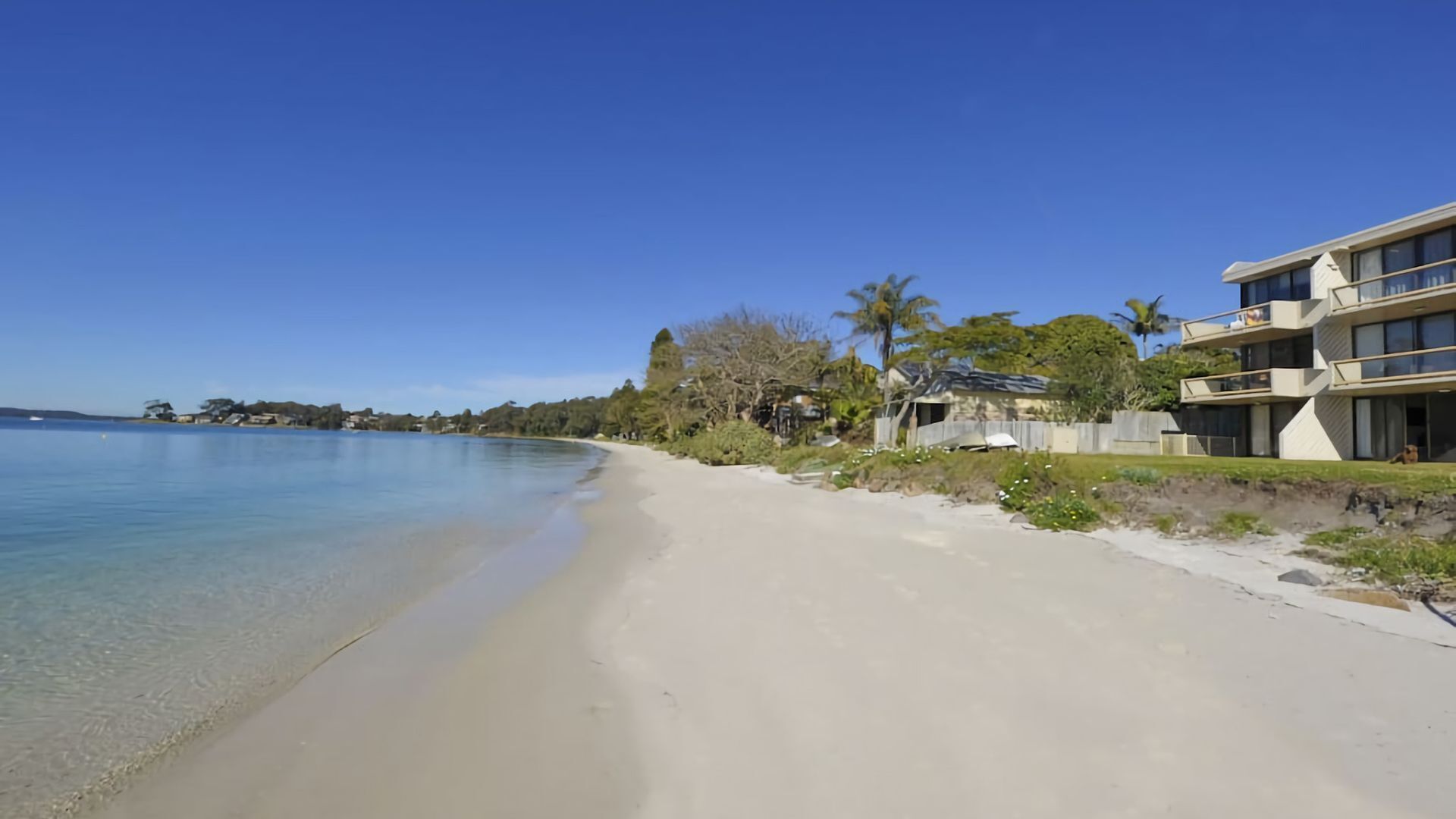 Une propriété située en bord de mer, avec une plage de sable, un ciel bleu dégagé et un bâtiment moderne doté de balcons.