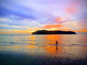 A beach scene with a person walking on the shore, calm water, and a sunset in the background.